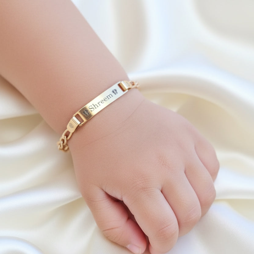 A gold-colored bracelet with the inscription 'Shreem' is held on a toddlers hand, showcasing its engraved design on a chain.