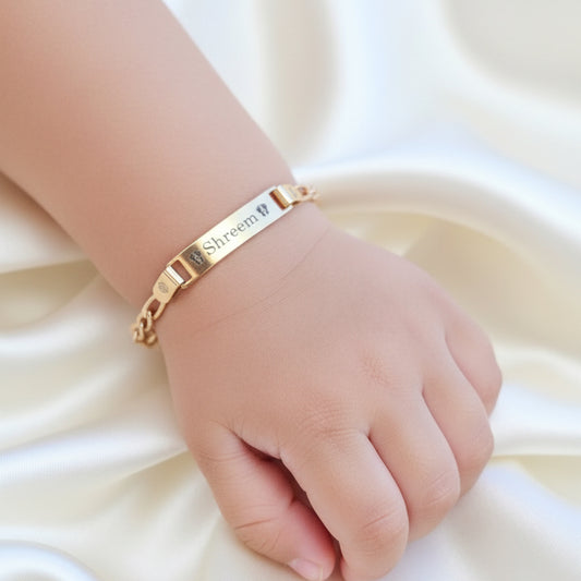 A gold-colored bracelet with the inscription 'Shreem' is held on a toddlers hand, showcasing its engraved design on a chain.