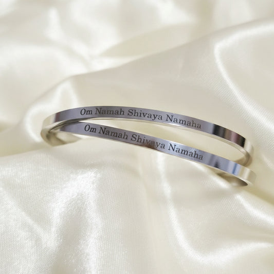 A stainless steel bracelet with the mantra 'Om Namah Shivaya Namaha' inscribed on it, displayed on a yellow fabric background.