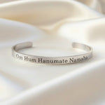 A silver-finished bracelet with the text 'Om Hanumate Namah' engraved on it, displayed on a textured background.