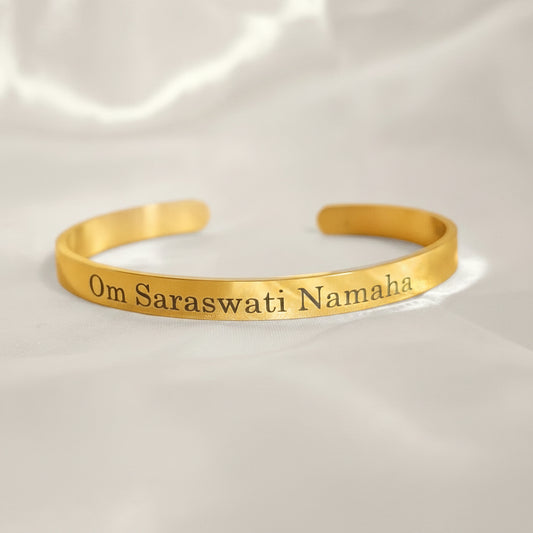 A gold-colored bracelet with the mantra 'Om Saraswati Namaha' engraved on it, displayed against a yellow background.